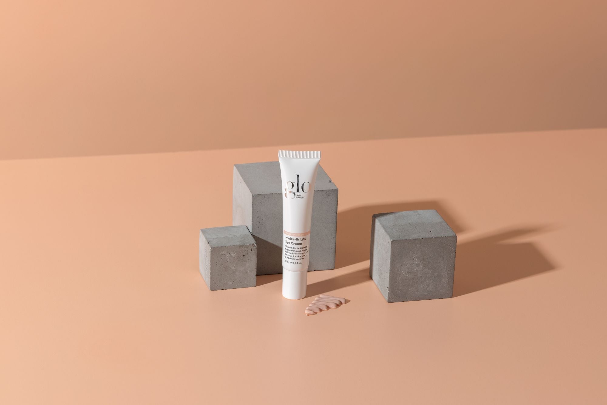 Tube of Glo Skin Beauty Hydra-Bright Eye Cream on peach background with concrete blocks and a cream swatch.