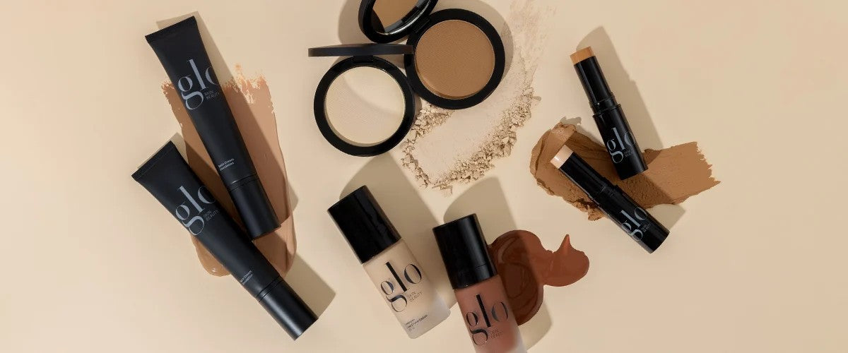 Assorted glo foundation bottles, tubes, compacts and sticks with foundation swatches on beige background
