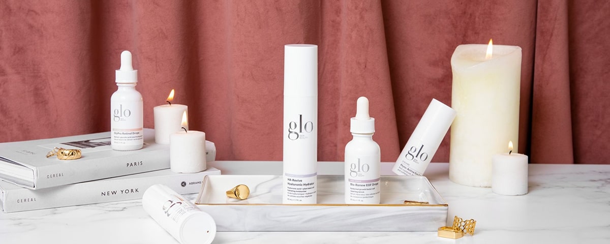 Front view of Glo Skin Beauty serums and dropper bottles showcasing white packaging and droppers on a marble tray with lit candles and stacked books