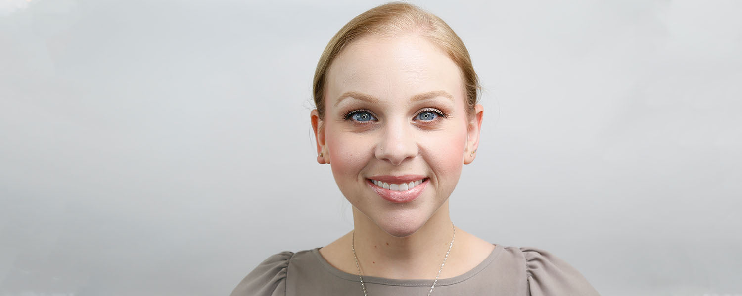 Smiling woman with light pink blush on the apples of her cheeks, center-parted hair pulled back, neutral gray background.