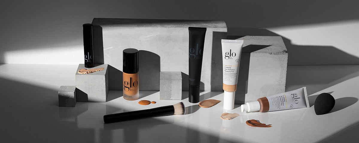 Studio view of Glo Skin Beauty foundation bottles and C-Shield Moisture Tint tube with brush, sponge, and swatches on a glossy tabletop