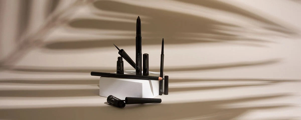 Various black eyeliners and pencil arranged on a white block with palm-leaf shadow backdrop
