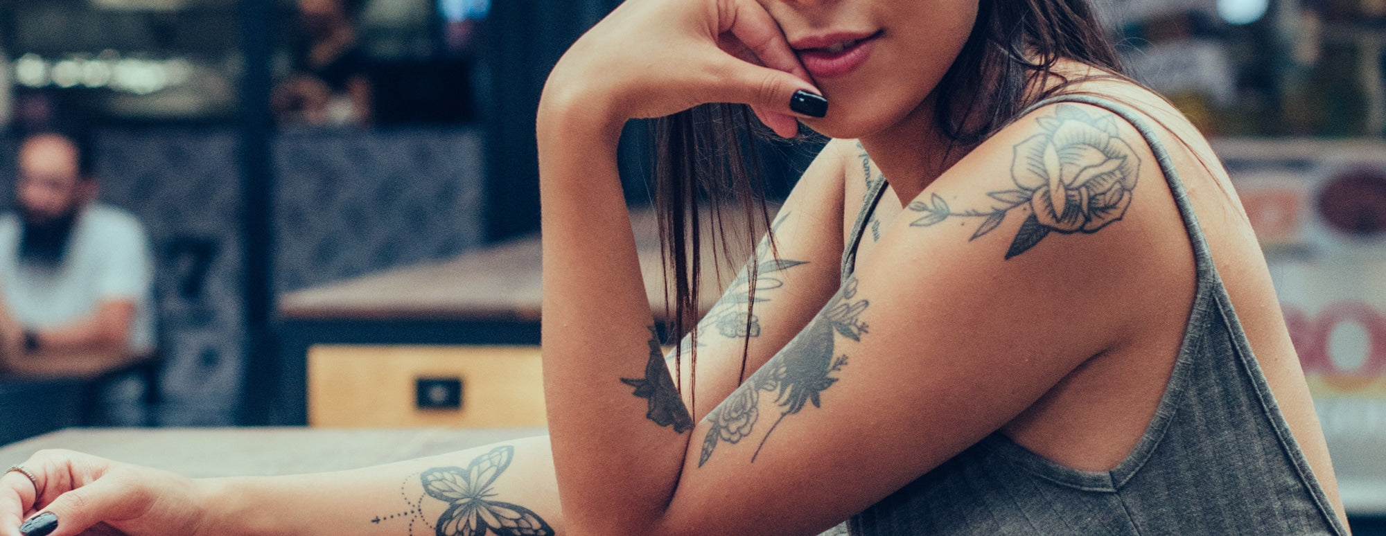 Young woman resting chin on hand, showing floral and butterfly tattoos on upper arm and forearm, gray tank top.
