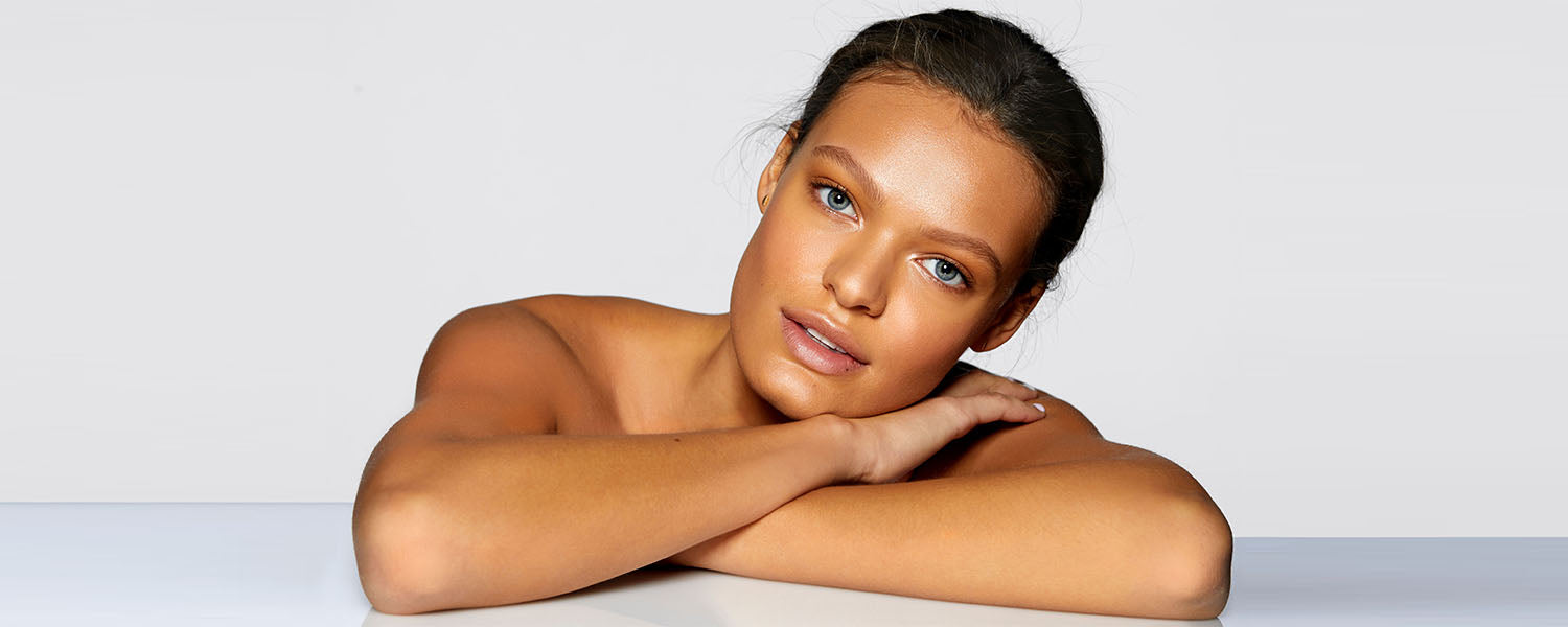 Young woman with glowing skin resting her chin on crossed forearms against a neutral gray background.