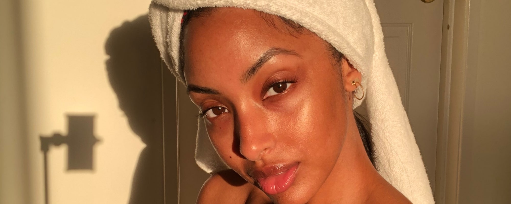 Close-up of a woman with towel-wrapped hair and glowing skin in warm sunlight, shadow of phone on wall.