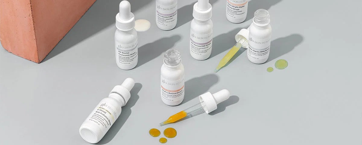 Assorted white dropper bottles of Glo Skin Beauty serums on gray surface with yellow and green serum drops