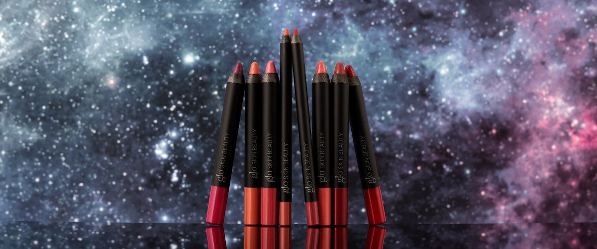 Assorted red and pink lip crayons with glo SKIN BEAUTY branding against a starry galaxy backdrop, reflected on glossy base.