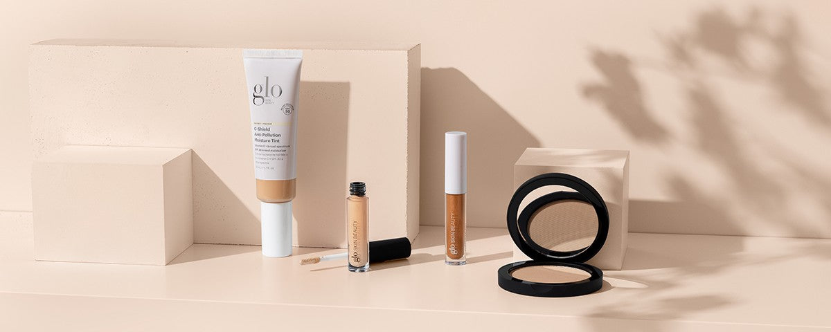Glo Skin Beauty makeup products on beige blocks: C-Shield Anti-Pollution Moisture Tint tube, two glosses, and compact powder