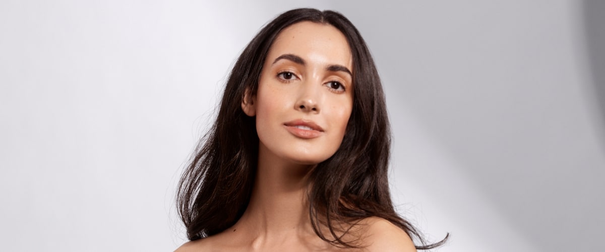 Woman with clear skin and long dark hair, bare shoulders, neutral expression in soft studio lighting