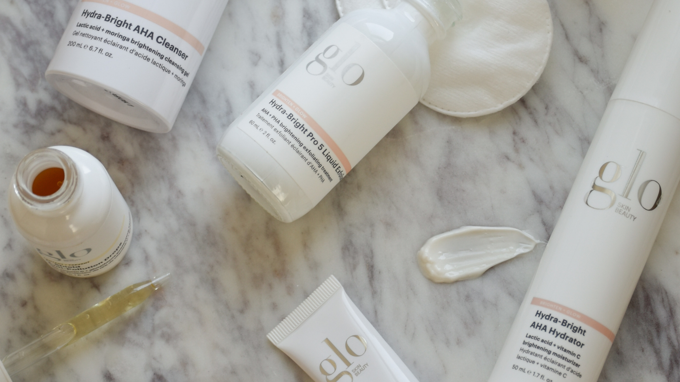 Flatlay of Glo Skin Beauty Hydra-Bright AHA Cleanser, Hydra-Bright Pro Liquid and AHA Hydrator bottles with cream on marble