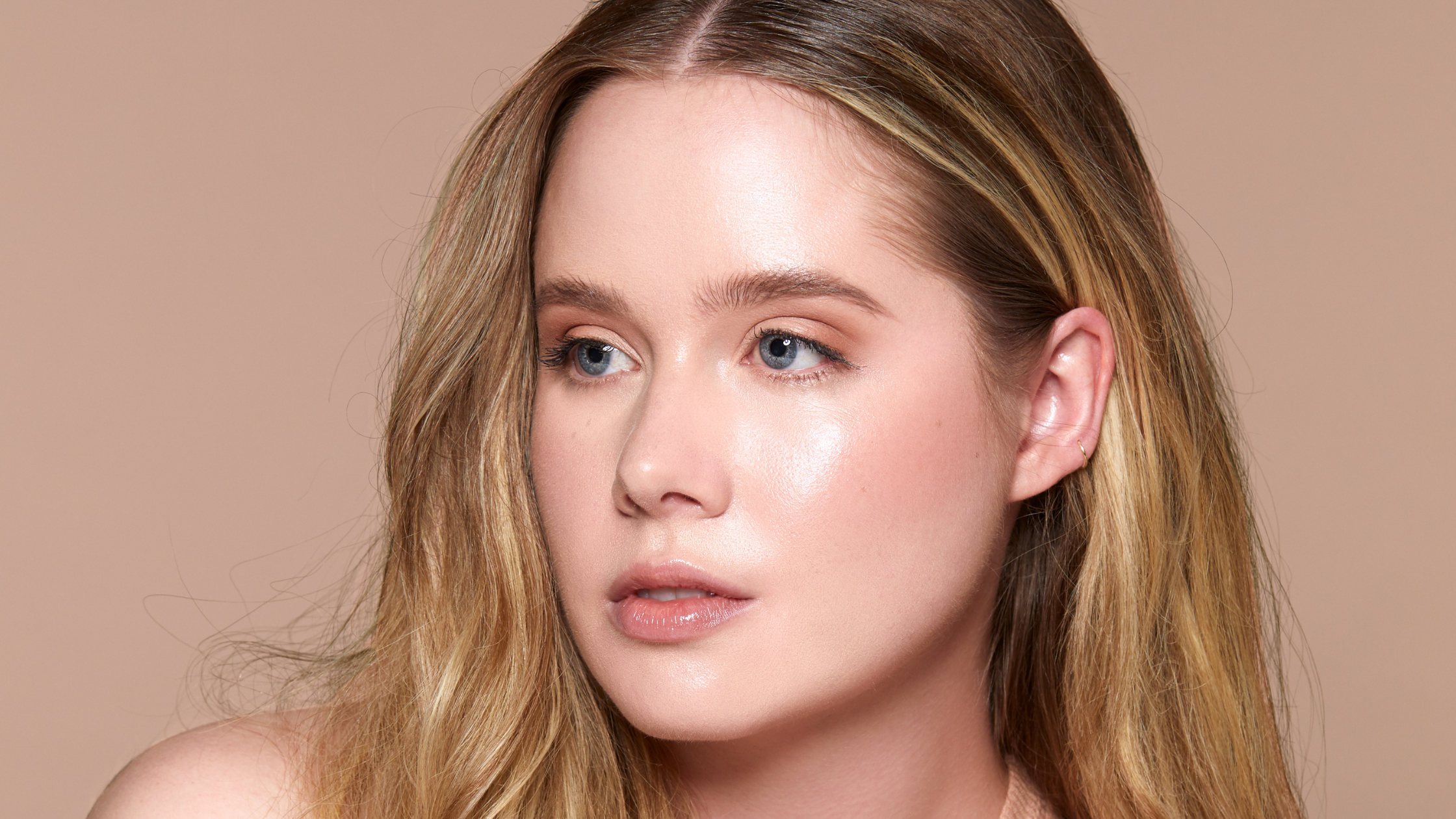 Close-up portrait of a young woman with dewy, glowing skin, blonde hair, and natural makeup against a neutral beige background.