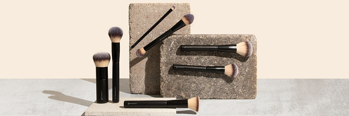 Seven black-handled Glo Skin Beauty makeup brushes arranged on and against textured concrete blocks on beige background