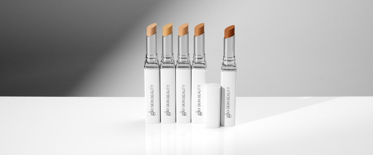 Five glo SKIN BEAUTY hybrid concealer sticks in graduated shades, white tubes standing upright