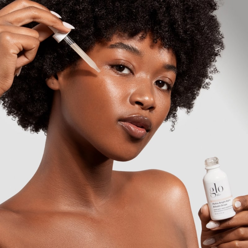 Woman applying drops from glo Hydra-Bright Alpha Arbutin Drops to her cheek, close-up showing bottle and dropper