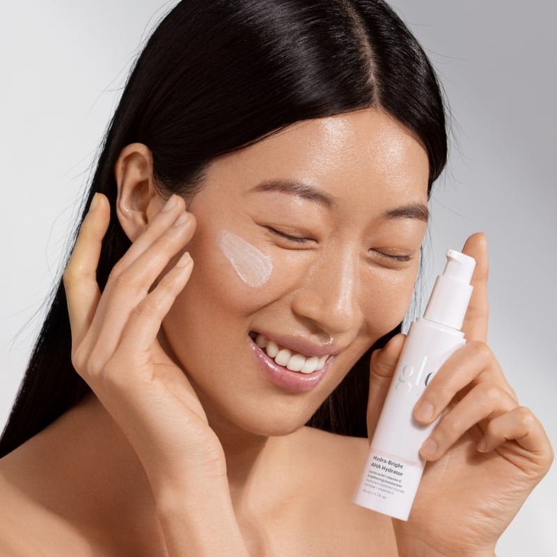 Smiling woman applying cream, holding bottle labeled glo Hydra-Bright AHA Hydrator.