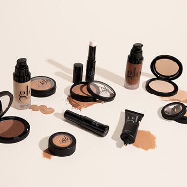 Assorted glo cosmetics: foundation pumps, compact powders, a stick concealer, and a cream tube arranged on a pale background