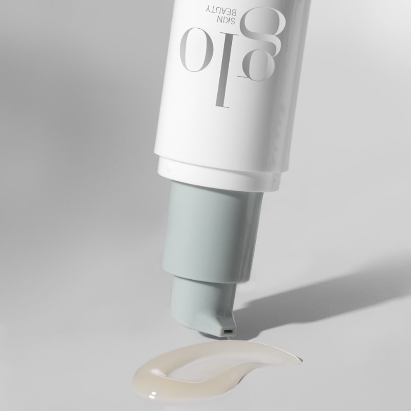 Upside-down white and pale-blue pump tube labeled glo SKIN BEAUTY dispensing a small dollop of cream onto a white surface.