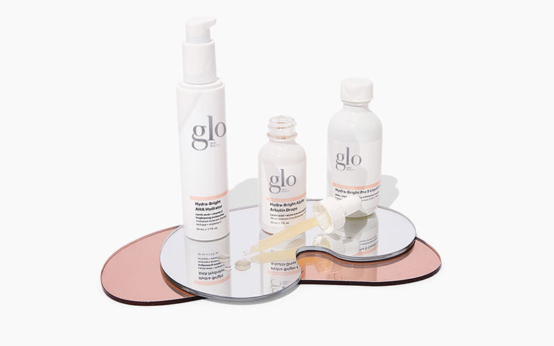 Three glo Hydra-Bright AHA products — Hydrator and Arbutin Drops on mirrored pink and silver trays