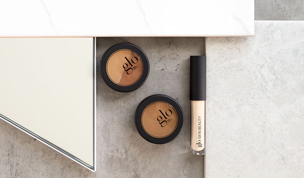 Two glo compact powders and a tube labeled 'glo SKIN BEAUTY' on a grey stone surface