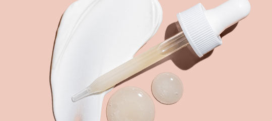 Glass dropper with skincare serum droplets and cream smear on peach background.