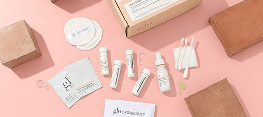 Assorted Glo Skin Beauty samples: vials, sachets and cotton pads arranged on pink background with glo SKIN BEAUTY card.