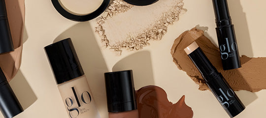 Assorted glo cosmetics with foundation sticks, bottles, powder and three foundation swatches in beige, tan and deep brown