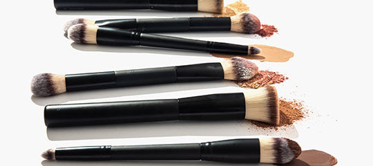 Six black-handled makeup brushes arranged horizontally with foundation and powder smudges on a white background.