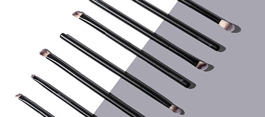 Nine black eye makeup brushes with angled and tapered bristles arranged diagonally on a two-tone gray background