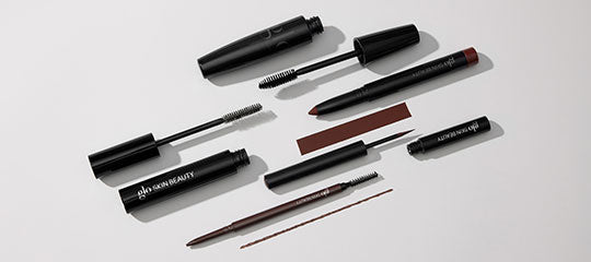 Assorted black mascara tubes, eyebrow pencils and spoolie brushes on a neutral background; branding 'glo SKIN BEAUTY' visible.