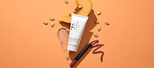 glo tube of skincare with powder, mauve lip-pencil strokes, and pumpkin slices and seeds on orange background