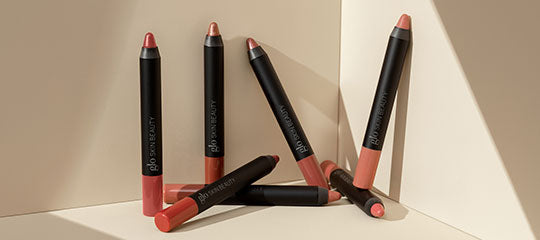 Six coral and nude lip crayons with black barrels arranged on a beige geometric background