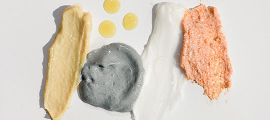 Four cosmetic product swatches (beige, charcoal gray, white, coral) and three yellow droplets on a white background.