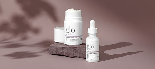 Two 'glo' skincare containers (jar and dropper bottle) on a stone platform against mauve background.