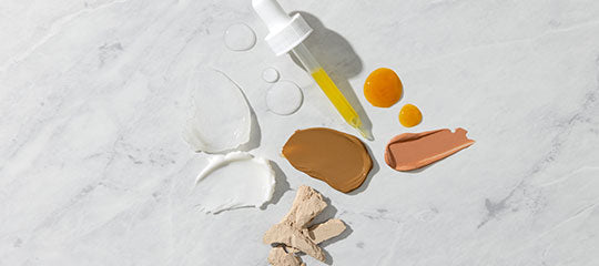 Dropper with yellow serum, droplets, cream and foundation swatches, and clay fragments on marble surface
