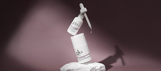 Two glo skincare products—a dropper bottle labeled glo and a cylindrical jar on a stone pedestal against a mauve background.