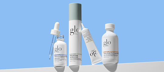 Four glo skincare containers: dropper bottle, tall pump, leaning tube, and capped bottle on white ledge, blue background.