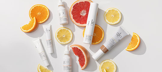 Assorted glo skincare tubes and serums arranged with sliced grapefruit, orange, and lemon on a white background.
