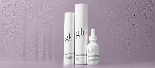 Three 'glo' skincare bottles—two tall cylinders and one dropper—on a mauve surface with water-droplet background.