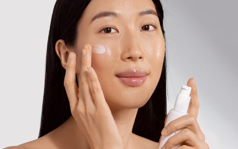 Woman applying moisturizer to her cheek while holding a white pump bottle