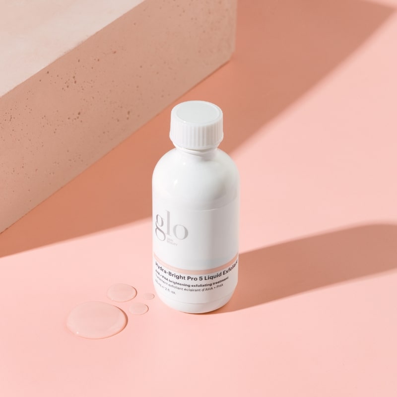 White "glo" bottle labeled "Hydra-Bright Pro 5 Liquid Exfoliant" with clear droplets on a pink surface
