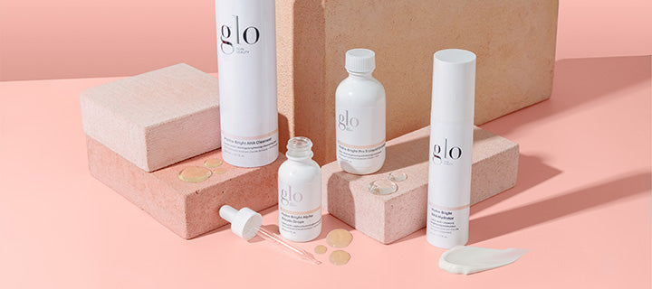 Three white 'glo' skincare bottles with dropper on pink blocks, serum drops and a cream smear.