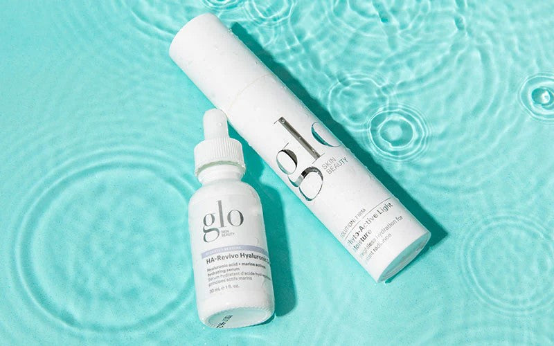 Two glo SKIN BEAUTY products on rippled aqua water: HA-Revive Hyaluronic dropper bottle and Hydra-Active tall bottle.