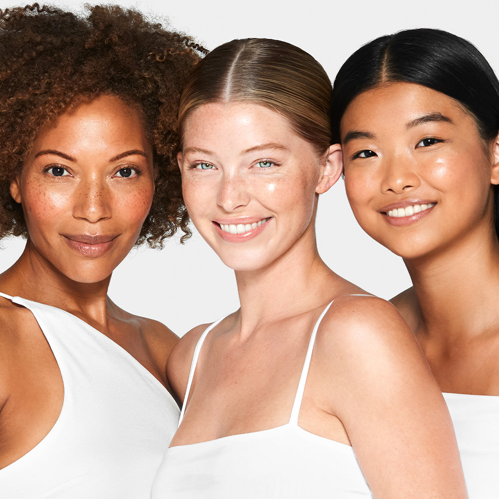 Three smiling women of different ethnicities in white tops, close-up portrait highlighting diverse skin tones.