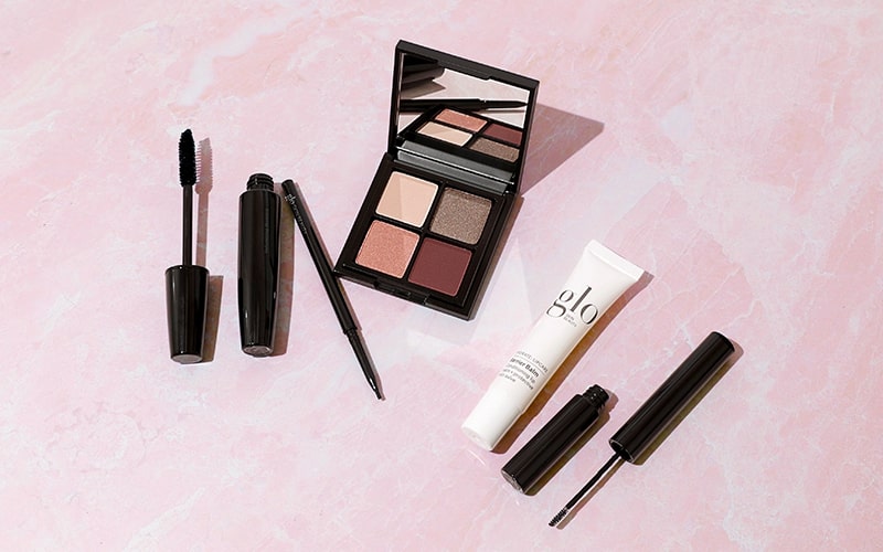 Eyeshadow quad, liquid eyeliner, two mascara wands and a white tube labeled 'glo' on pink marble surface.
