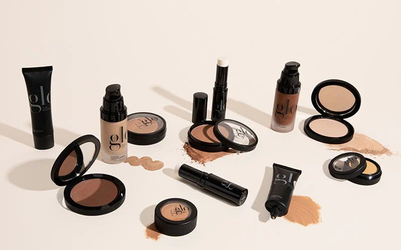 Assorted glo cosmetics: liquid foundations, cream stick, compacts and powder swatches on a beige background