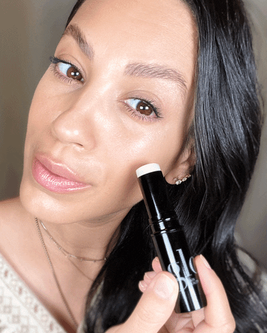 Woman with dewy makeup holding a black cream stick near her cheek, showing product application on skin