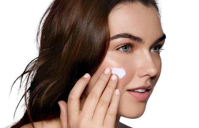 Woman applying white facial cream to her right cheek with fingertips, close-up of face.