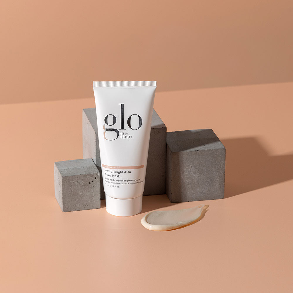 Tube of glo SKIN BEAUTY Hydra-Bright AHA Glow Mask on peach background with gray concrete blocks and a cream smear.