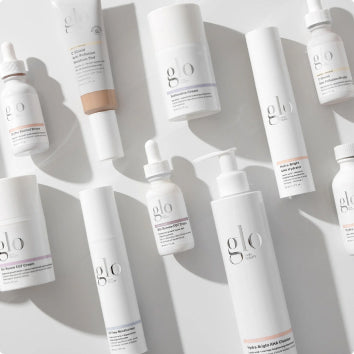 Flat lay of white glo skincare bottles and tubes arranged on a pale surface with soft shadows