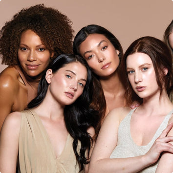 Four women of diverse skin tones posing closely against a neutral background, showcasing natural makeup and dewy skin.